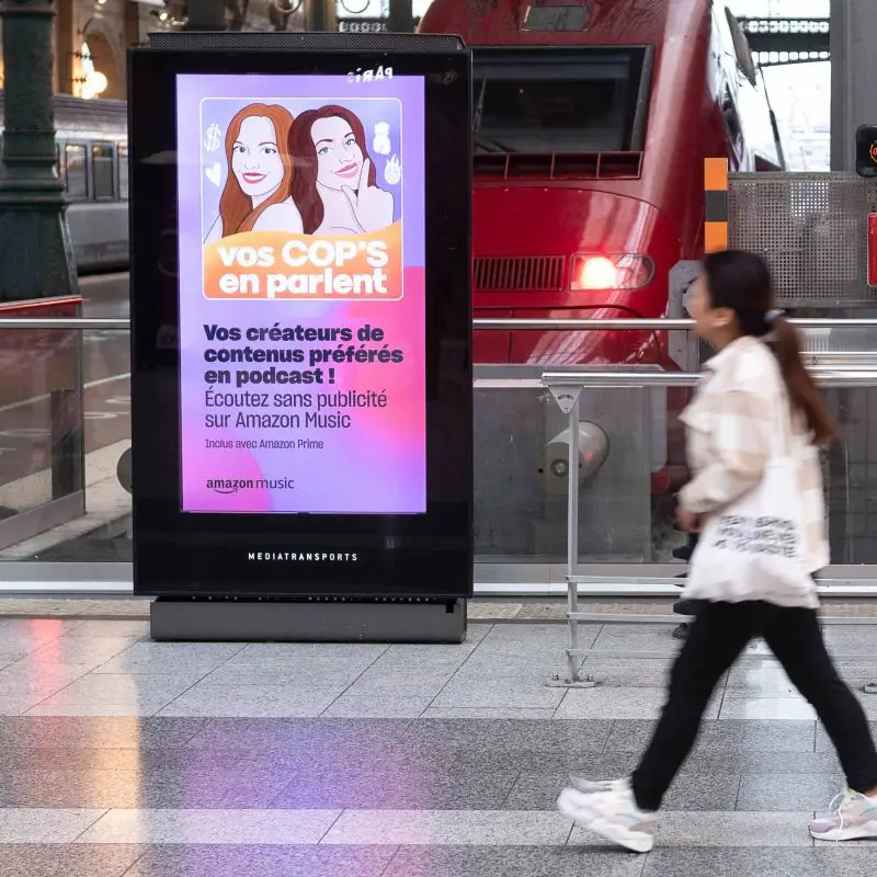 Affiche publicitaire du podcast Vos Cop's en Parlent sur un panneau numérique en gare, campagne Amazon Music avec les deux créatrices illustrées, par La Renarde Bouclée